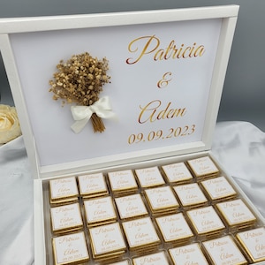 Op de afbeelding: Een witte doos met een gouden inscriptie die "Patricia & Adem 09.09.2023" luidt. De doos is open en bevat 25 kleine witte vierkanten met gouden letters die "Patricia & Adem 09.09.2023" luiden.