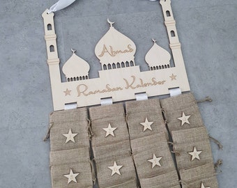 May include: Wooden Ramadan calendar shaped like a mosque with 12 burlap bags hanging from it. The bags are numbered 1 through 12 and have a star on each. The calendar says "Abnas Ramadan Kalender" on the top.
