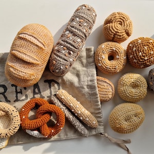 Könnte beinhalten: Eine Sammlung gehäkelter Brot- und Gebäckspielzeuge in verschiedenen Farben und Formen, darunter ein Laib Brot, ein Brezel, ein Bagel und ein Donut. Die Spielzeuge sind auf einer beigefarbenen Stofftasche mit dem Wort "Brot" darauf angeordnet.