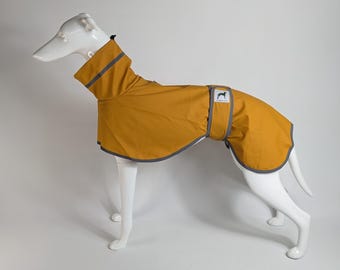 May include: A mustard yellow dog raincoat featuring a high collar, grey reflective trim along the edges, and a belly strap with a white rectangular label displaying a black silhouette of a greyhound and the text Greyhound Gear.