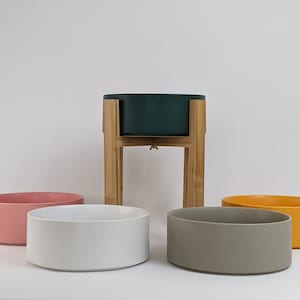 May include: A selection of ceramic pet bowls in shades of pink, white, grey, and yellow. One bowl is elevated on a wooden stand. The bowls are cylindrical and have a minimalist design, suitable for pet food or water.
