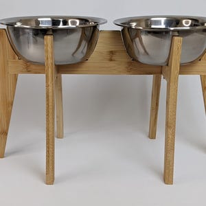 May include: A raised pet feeder featuring two removable stainless steel bowls set into a light-coloured bamboo wood stand. The stand has four angled legs and a horizontal support frame that holds the bowls securely above the ground surface.
