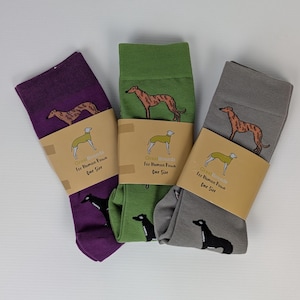 May include: Three pairs of crew socks in purple, green, and grey, each featuring a repeating pattern of brown and black greyhound dogs. Each pair is secured with a tan paper band printed with a dog illustration and the text Great Hounds For Human Paws One Size.