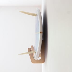 Peut inclure: Une planche de surf avec un motif marbré bleu clair et blanc est montée verticalement sur un mur à l'aide d'un support en bois clair. La planche de surf présente deux ailerons de couleur claire s'étendant sur le côté, fixés dans le support.