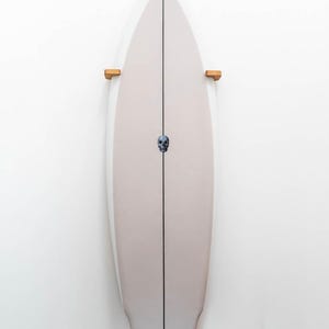 May include: A surfboard with a light tan deck and a black stripe down the center. A small skull graphic is centered on the board. The board is mounted on two wooden brackets against a white wall.