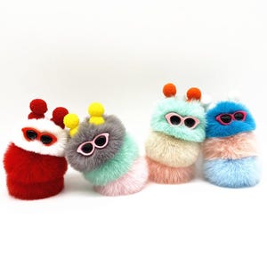 Pode incluir: Quatro brinquedos de pelúcia empilhados e coloridos que se assemelham a lagartas, cada um com óculos de sol e uma antena de pompom. Os brinquedos são vermelhos, cinza, verde menta e azul, com detalhes em rosa, amarelo e laranja. Em um fundo branco.