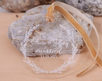 Pode incluir: Enfeite de casamento em acrílico transparente com design de coroa de flores branca. Os nomes "Olivia & Jeffery" e a palavra "casados" estão inscritos, juntamente com a data "12.19.2021". Uma fita de veludo bege está presa para pendurar.