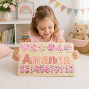 May include: A rectangular wooden puzzle board featuring the name Amanda in large pink letters. The top row includes six shape cutouts labeled heart, square, star, circle, triangle, and hexagon. The bottom row displays numbers one through ten.
