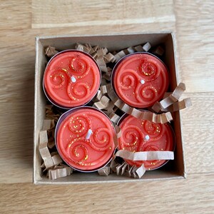 May include: Four orange scented tea lights in a cardboard box with brown paper packing. Each tea light has a white wick and a gold glitter design.