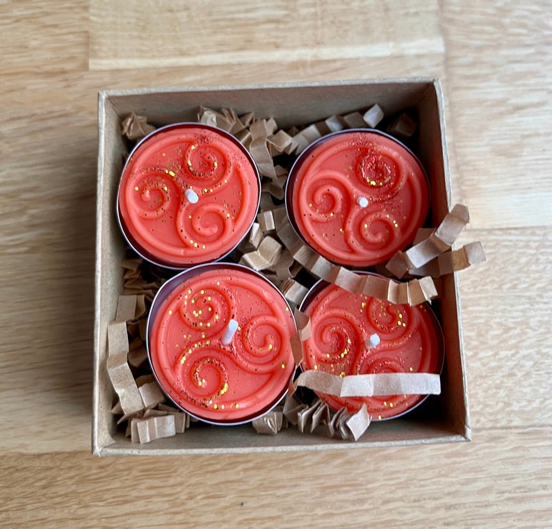 May include: Four orange scented tea lights in a cardboard box with brown paper packing. Each tea light has a white wick and a gold glitter design.