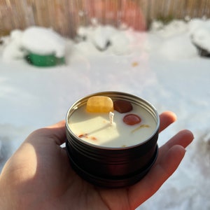May include: A small, black tin candle with a white wick and embedded crystals. The crystals are orange, red, and yellow. The candle is sitting on a hand.