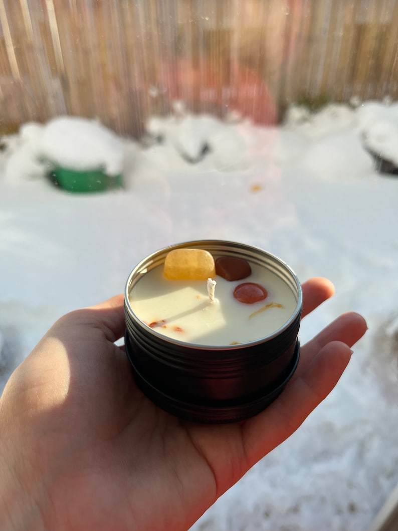 May include: A small, black tin candle with a white wick and embedded crystals. The crystals are orange, red, and yellow. The candle is sitting on a hand.
