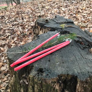 May include: Three long, bright pink candles are arranged on a tree stump in a woodland setting. The candles are lying on top of a small evergreen branch.
