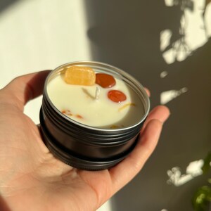 May include: A small black tin candle with a white wax top. The candle has three small, colorful stones embedded in the wax.