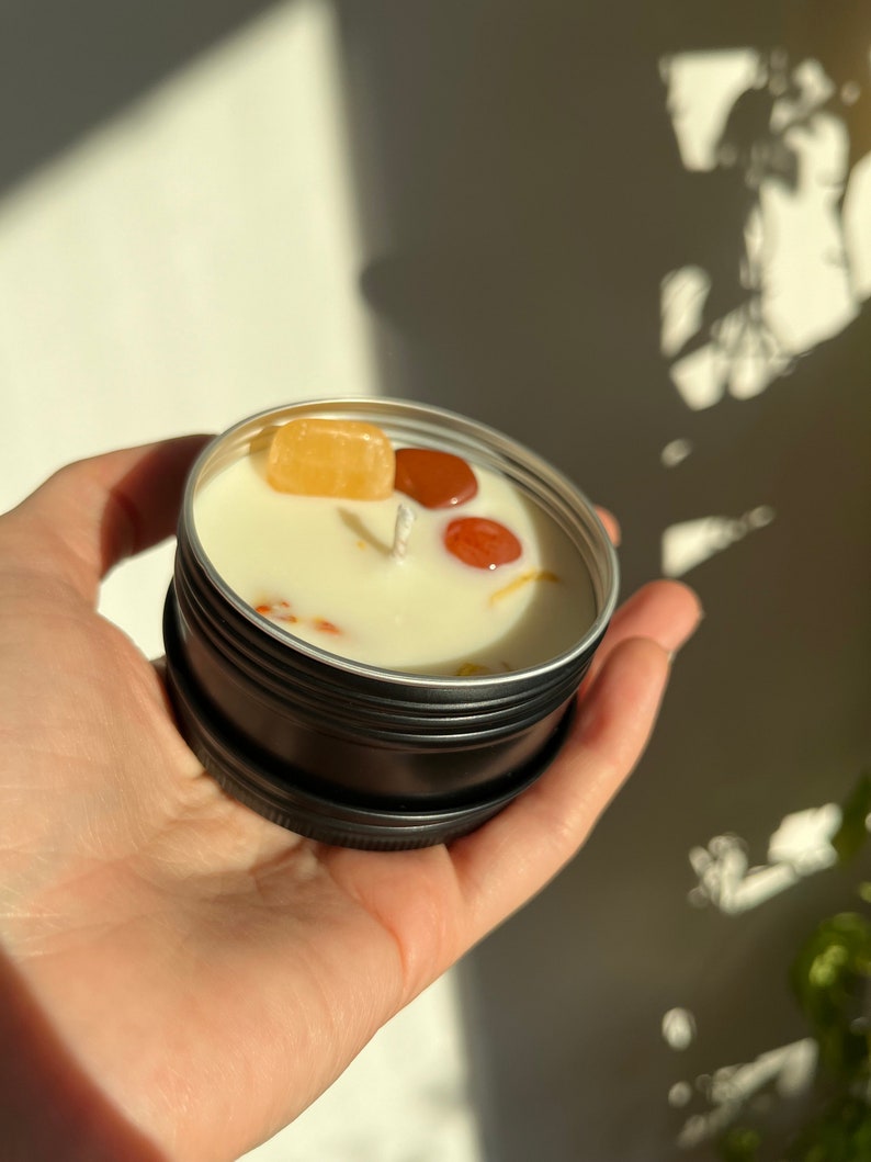 May include: A small black tin candle with a white wax top. The candle has three small, colorful stones embedded in the wax.