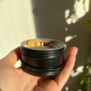 May include: A small black metal tin candle with a yellow and brown crystal on top. The candle is in a person's hand.