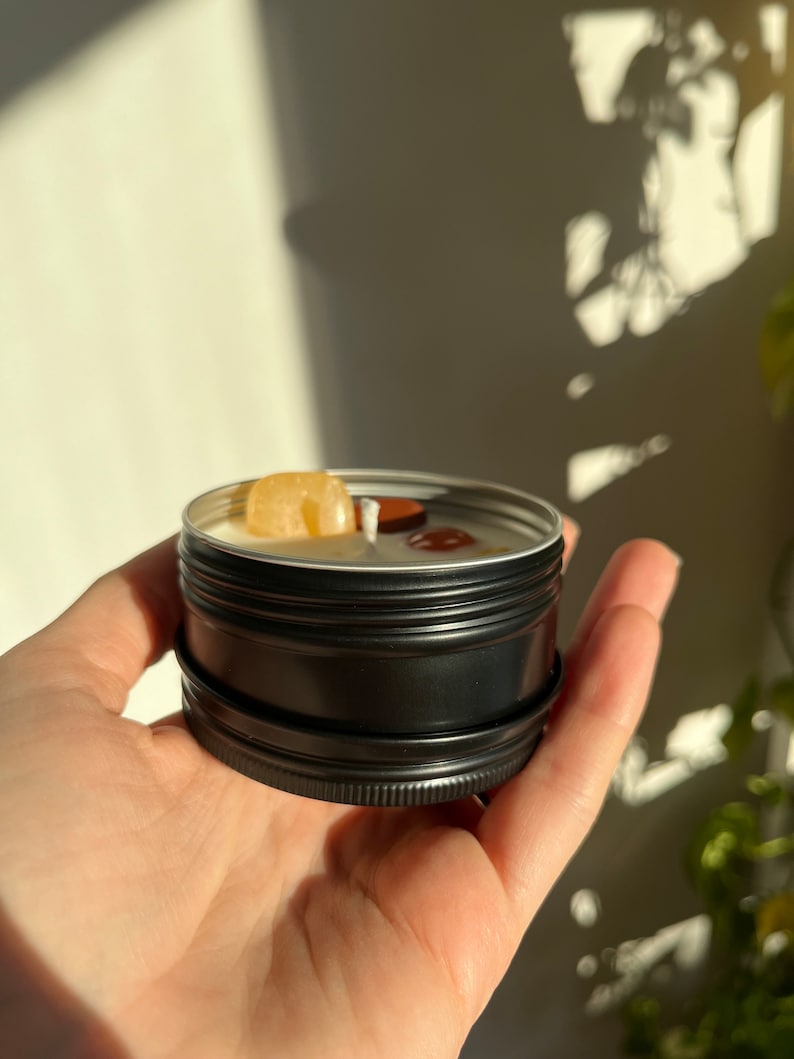 May include: A small black metal tin candle with a yellow and brown crystal on top. The candle is in a person's hand.