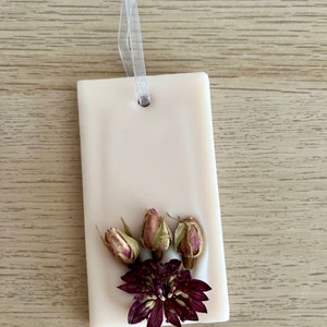 Puede incluir: Un derretido de cera rectangular blanco con un agujero en la parte superior para colgar. El derretido de cera est&aacute; decorado con flores secas, incluyendo tres peque&ntilde;os capullos de rosa y una flor morada m&aacute;s grande.
