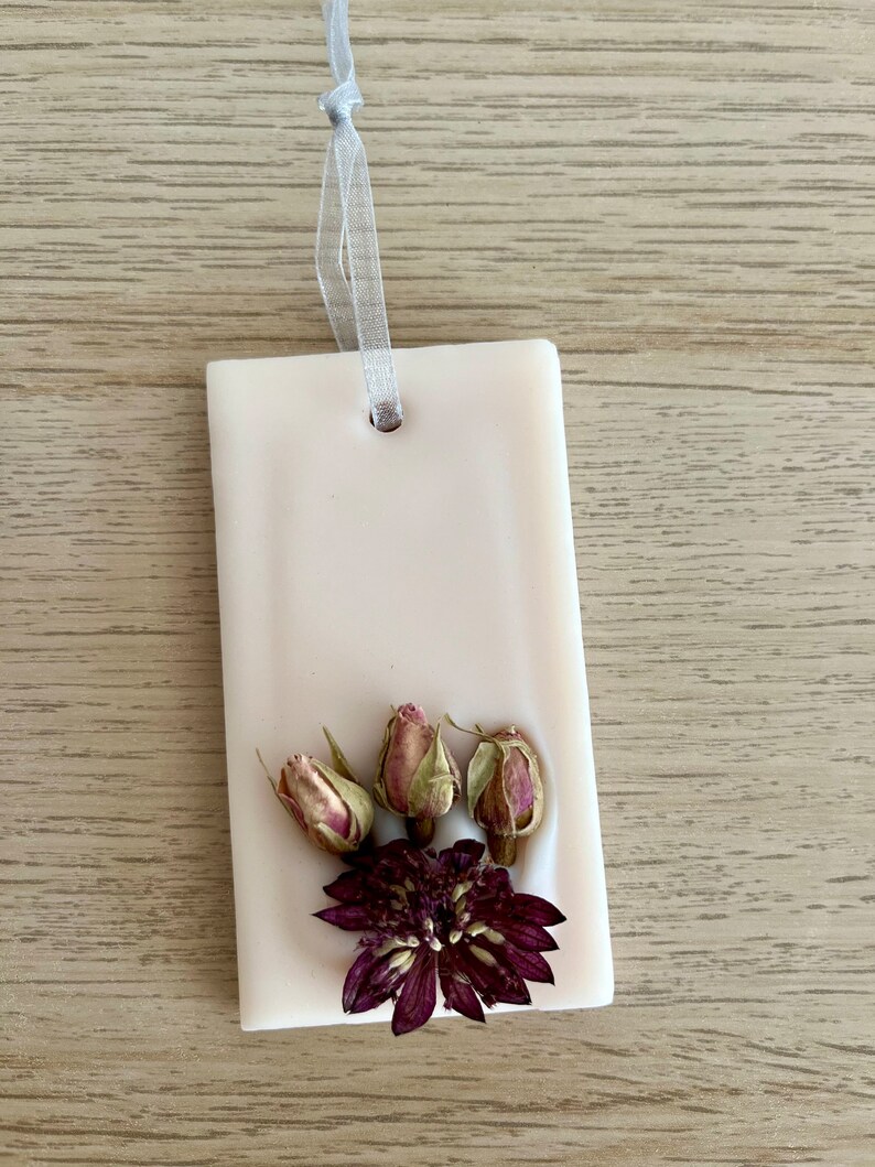 Puede incluir: Un derretido de cera rectangular blanco con un agujero en la parte superior para colgar. El derretido de cera est&aacute; decorado con flores secas, incluyendo tres peque&ntilde;os capullos de rosa y una flor morada m&aacute;s grande.