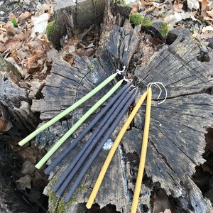 May include: A group of six long, thin candles in three different colours: yellow, green, and black. The candles are arranged in a circle on a tree stump.