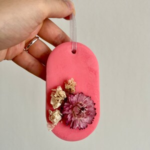 Puede incluir: Un derretido de cera de forma ovalada rosa con flores secas incrustadas en la cera. El derretido tiene una cinta blanca unida a la parte superior para colgar.