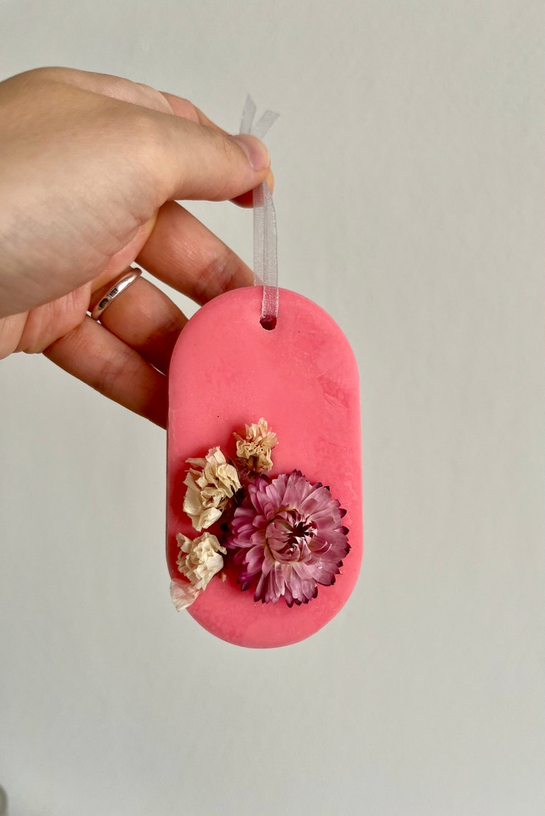 Puede incluir: Un derretido de cera de forma ovalada rosa con flores secas incrustadas en la cera. El derretido tiene una cinta blanca unida a la parte superior para colgar.