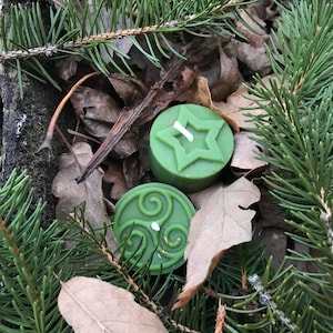 Op de afbeelding: Twee groene geurkaarsen met een ster- en een driedubbele spiraalvorm. De kaarsen liggen op een bed van dennennaalden en bladeren.