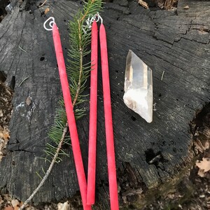 May include: Three long, thin, red candles with white wicks on a tree stump. A clear quartz crystal is also on the stump.