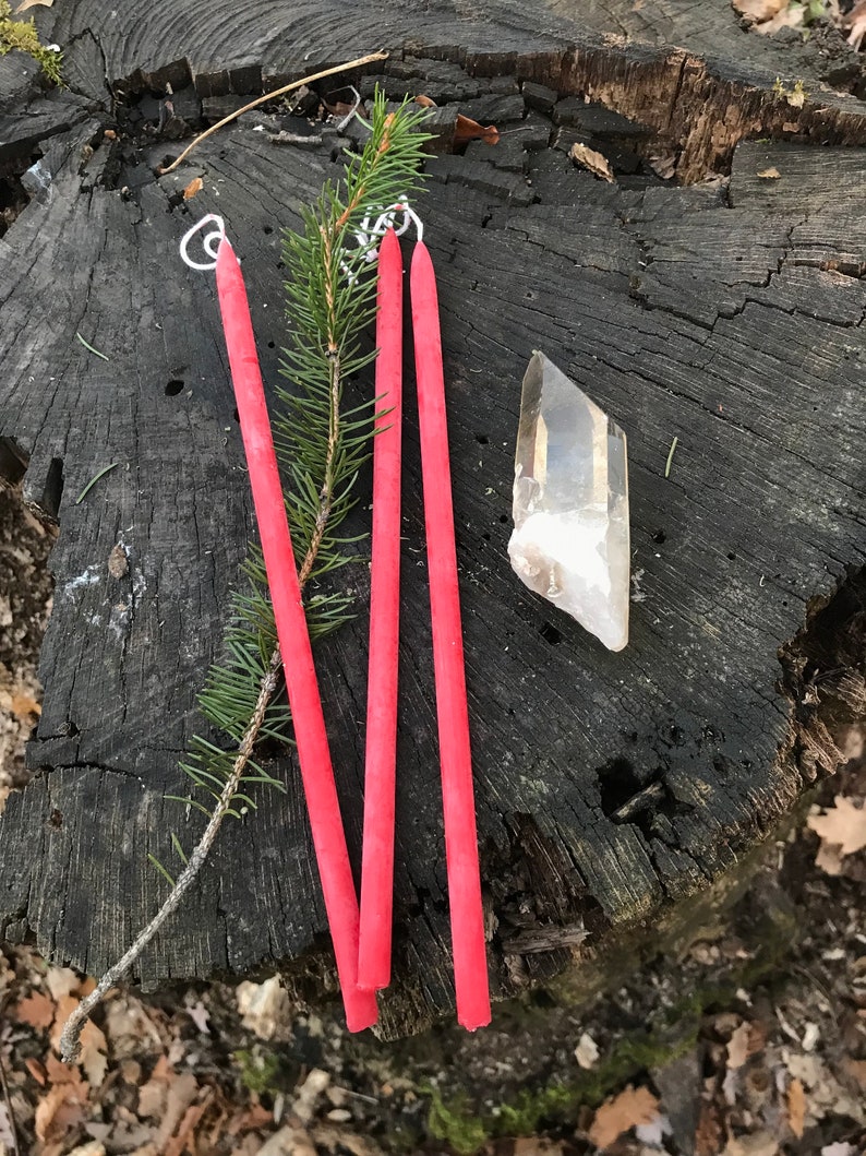 May include: Three long, thin, red candles with white wicks on a tree stump. A clear quartz crystal is also on the stump.