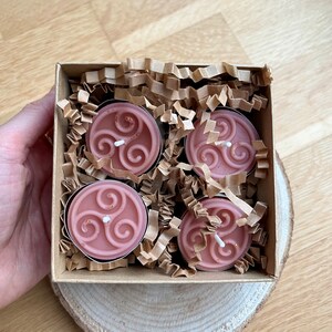 May include: Four pink tea light candles in a cardboard box with brown paper packing material. Each candle has a white wax design of a triple spiral.
