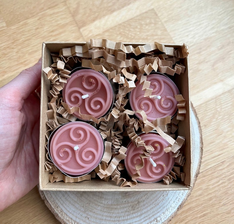 May include: Four pink tea light candles in a cardboard box with brown paper packing material. Each candle has a white wax design of a triple spiral.