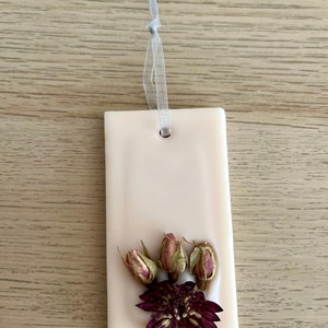 Puede incluir: Un adorno colgante de cer&aacute;mica blanca con un arreglo de flores secas. El arreglo incluye una flor morada y tres capullos de rosa secos.