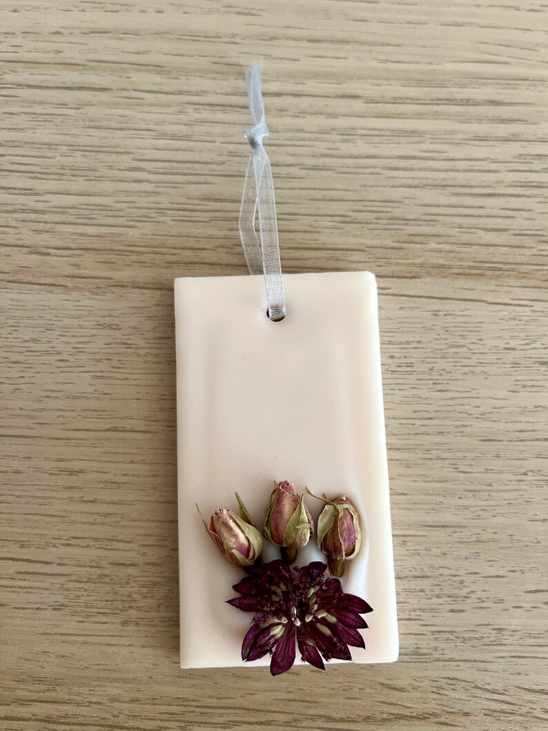 Puede incluir: Un adorno colgante de cer&aacute;mica blanca con un arreglo de flores secas. El arreglo incluye una flor morada y tres capullos de rosa secos.