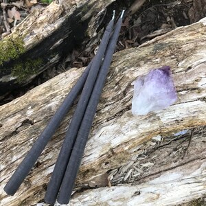 May include: Three black taper candles and a purple amethyst crystal rest on a weathered log. The candles are slender and have white wicks. The amethyst is a light purple color and has a rough, natural texture. The log has a mix of light and dark wood.
