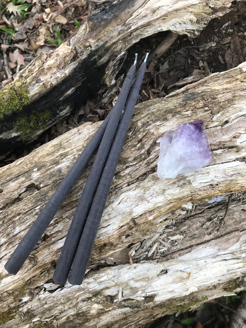 May include: Three black taper candles and a purple amethyst crystal rest on a weathered log. The candles are slender and have white wicks. The amethyst is a light purple color and has a rough, natural texture. The log has a mix of light and dark wood.