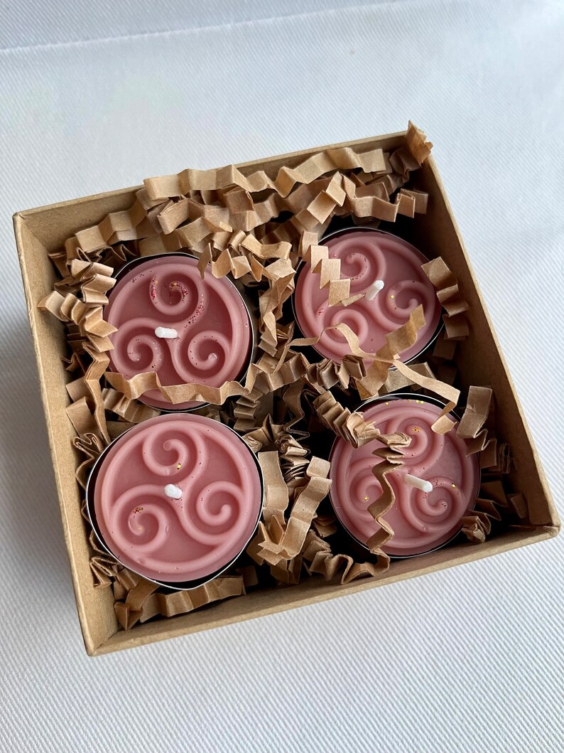 May include: Four pink tea light candles with a gold triquetra design in a brown cardboard box with shredded paper packing.