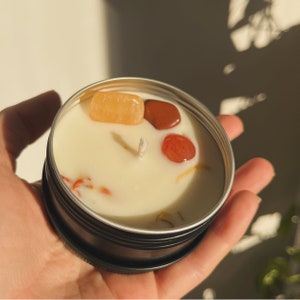 May include: A small, round candle in a black tin container with a white wax base. The candle has a white wick and is decorated with small, colorful stones.