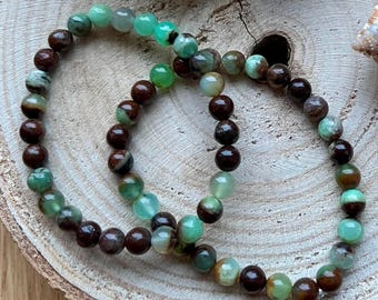 Puede incluir: Dos pulseras hechas con cuentas de piedras preciosas verdes y marrones. Las pulseras están apiladas una encima de la otra sobre una superficie de madera.