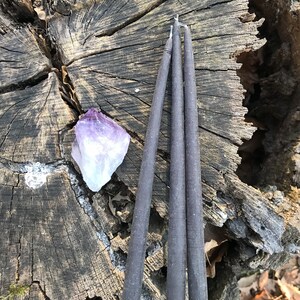 May include: Three dark gray taper candles are arranged on a weathered tree stump, next to a purple amethyst crystal. The candles are slender and have a rustic appearance. The background includes wood and foliage.