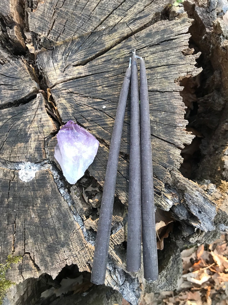 May include: Three dark gray taper candles are arranged on a weathered tree stump, next to a purple amethyst crystal. The candles are slender and have a rustic appearance. The background includes wood and foliage.