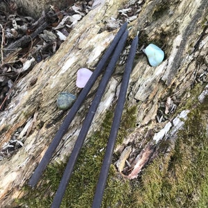 May include: Three black incense sticks on a mossy log with two small stones, one pink and one blue-green.