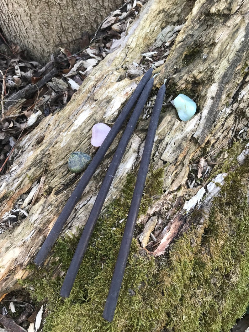 May include: Three black incense sticks on a mossy log with two small stones, one pink and one blue-green.