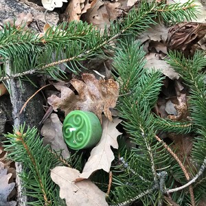 Op de afbeelding: Een groene kaars met een Keltisch knoopontwerp erop, omringd door bruine bladeren en groene dennennaalden.