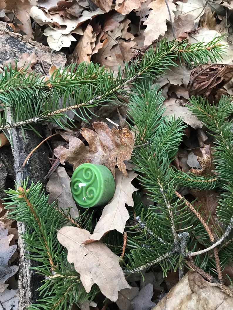 Op de afbeelding: Een groene kaars met een Keltisch knoopontwerp erop, omringd door bruine bladeren en groene dennennaalden.