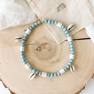 May include: A light blue and white beaded necklace with small white seashells hanging from it. The necklace is on a wooden surface and a small white bag with the text "Naturstein Calcit T&uuml;rkis" and "Aloha Dogs" is in the background.