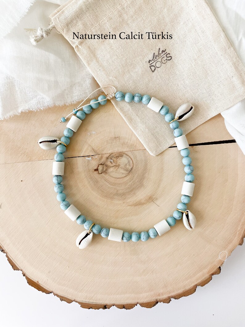 May include: A light blue and white beaded necklace with small white seashells hanging from it. The necklace is on a wooden surface and a small white bag with the text "Naturstein Calcit T&uuml;rkis" and "Aloha Dogs" is in the background.