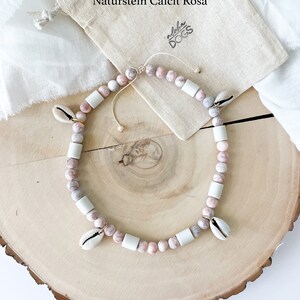 May include: A pink and white beaded necklace with shell charms. The necklace is on a wooden surface and a small white bag with the text "Aloha Dogs" is in the background. The text "Naturstein Calcit Rosa" is also visible in the background.