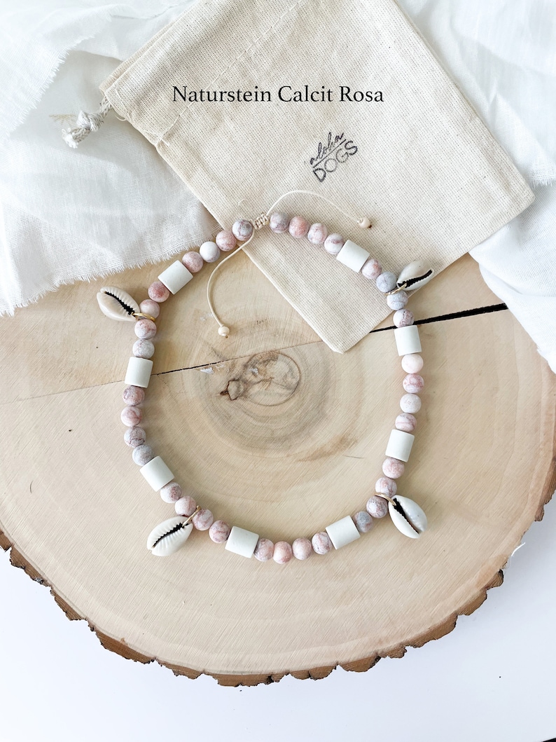May include: A pink and white beaded necklace with shell charms. The necklace is on a wooden surface and a small white bag with the text "Aloha Dogs" is in the background. The text "Naturstein Calcit Rosa" is also visible in the background.