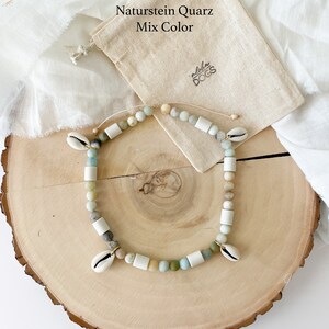 May include: A natural stone quartz necklace with a mix of colors, including white, green, and brown. The necklace is made with small, round beads and features two small seashells as accents. The necklace is displayed on a wooden surface with a small, beige drawstring bag.