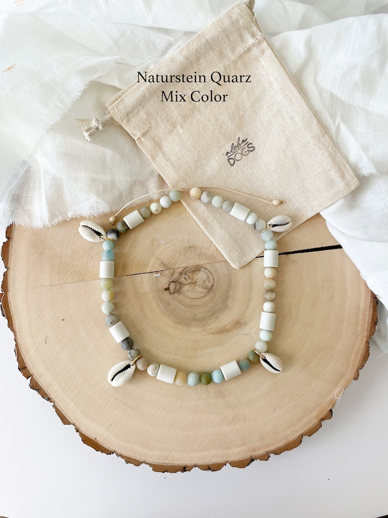 May include: A natural stone quartz necklace with a mix of colors, including white, green, and brown. The necklace is made with small, round beads and features two small seashells as accents. The necklace is displayed on a wooden surface with a small, beige drawstring bag.